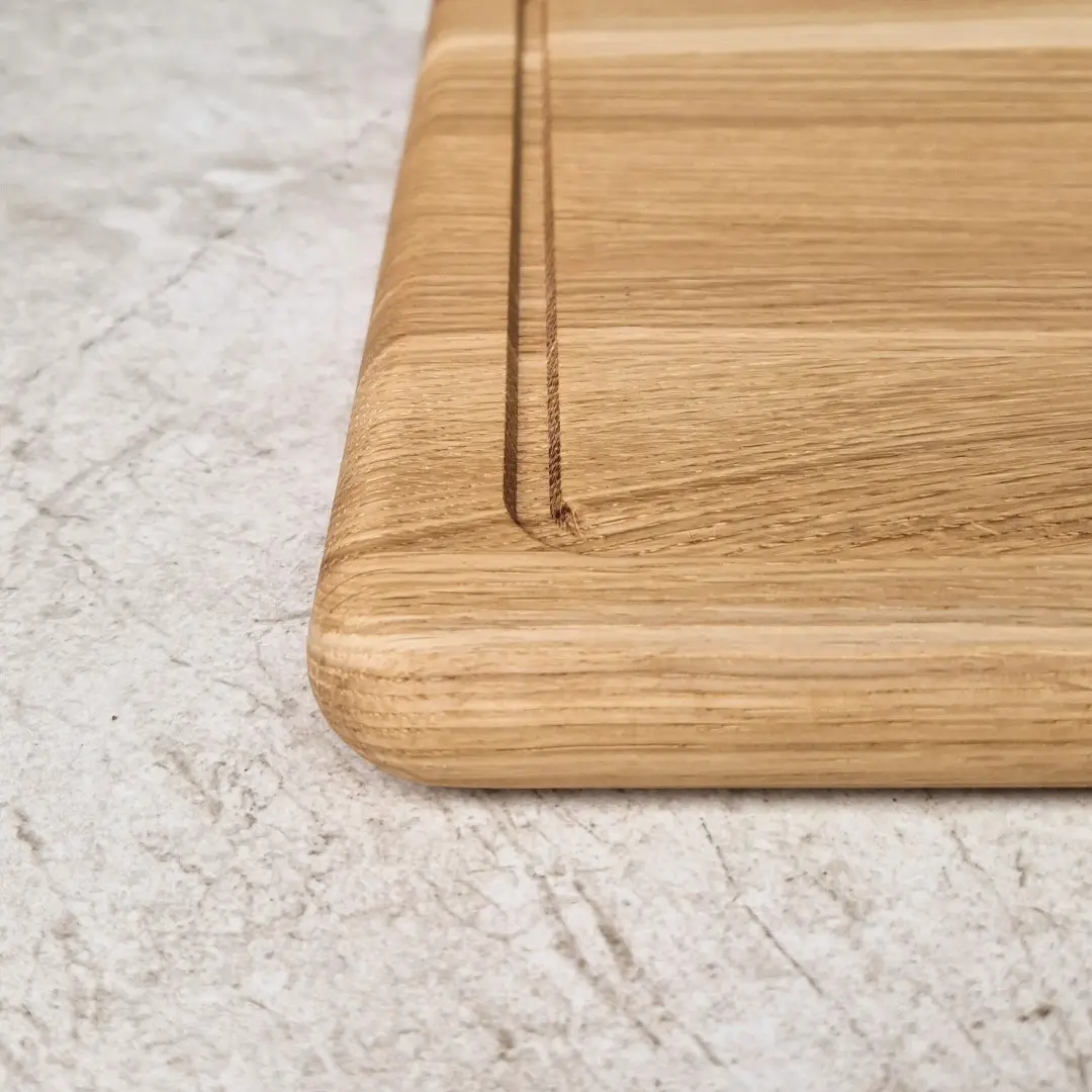 Zaokrąglony róg grubej gładkiej jasnej deski z rowkiem wykończonej twardym woskiem, masywna jasna deska do serwowania, deska do krojenia z łagodnymi  krawędziami, polski producent grubych desek dębowych i jesionowych, ekologiczna deska do krojenia, bezpieczna deska do krojenia, Rounded corner of a thick, smooth, light-colored board with a groove, finished with hard wax, solid light-colored serving board, cutting board with smooth edges, Polish manufacturer of thick oak and ash boards, eco-friendly cutting board, safe cutting board